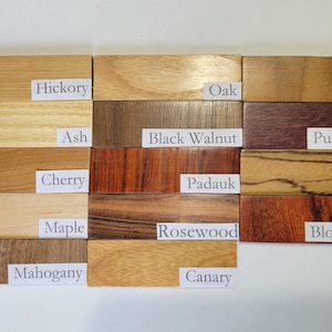 Pu&ograve; includere: Primo piano di 15 campioni di legno, ognuno etichettato con il suo nome. I campioni di legno sono disposti in tre file di cinque. I campioni di legno sono tutti di colori e texture diversi. I campioni di legno sono etichettati come segue: Hickory, Oak, Teak, Ash, Black Walnut, Purpleheart, Cherry, Padauk, Zebra, Maple, Rosewood, Bloodwood, Mahogany, Canary.