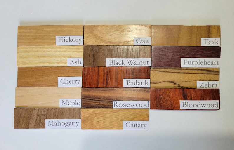 Pu&ograve; includere: Primo piano di 15 campioni di legno, ognuno etichettato con il suo nome. I campioni di legno sono disposti in tre file di cinque. I campioni di legno sono tutti di colori e texture diversi. I campioni di legno sono etichettati come segue: Hickory, Oak, Teak, Ash, Black Walnut, Purpleheart, Cherry, Padauk, Zebra, Maple, Rosewood, Bloodwood, Mahogany, Canary.