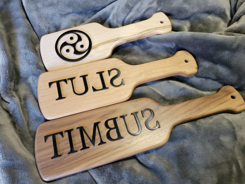 Pu&ograve; includere: Tre palette di legno con diversi disegni. La paletta superiore ha un simbolo triquetra nero. La paletta centrale ha la parola "SLUT" incisa su di essa. La paletta inferiore ha la parola "SUBMIT" incisa su di essa.
