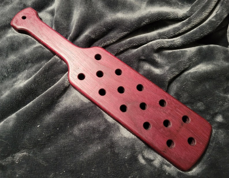 May include: A wooden paddle with a dark red stain and multiple holes drilled into the surface.