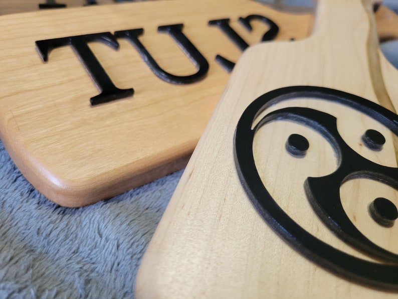 Pu&ograve; includere: Due palette di legno con disegni dipinti di nero. Una paletta ha la parola "TUZ" su di essa. L'altra paletta ha un cerchio nero con un design stilizzato al suo interno.