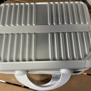 May include: A rectangular silver hard-shell travel case featuring a textured finish with vertical ribbed detailing. The case includes a wide horizontal elastic band across the center and a sturdy, rounded handle attached to the bottom edge.