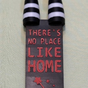 May include: A decorative bookmark featuring the legs of the Wicked Witch of the East from The Wizard of Oz. The legs are black and white striped with red shoes. The bookmark has the text "There's no place like home".