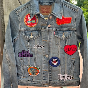 May include: A light blue denim jacket with various band patches, including "Peace Love Music Woodstock", "Jimi Hendrix", "The Who", "Elvis", "Dowie", "Allman Brothers Band", "Doors", and "Bon Jovi".