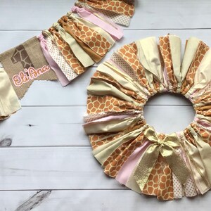 May include: A brown, pink, and gold fabric garland with a giraffe print and the text "Eli Ana" on a burlap banner. The garland is made of multiple strips of fabric with a frayed edge.