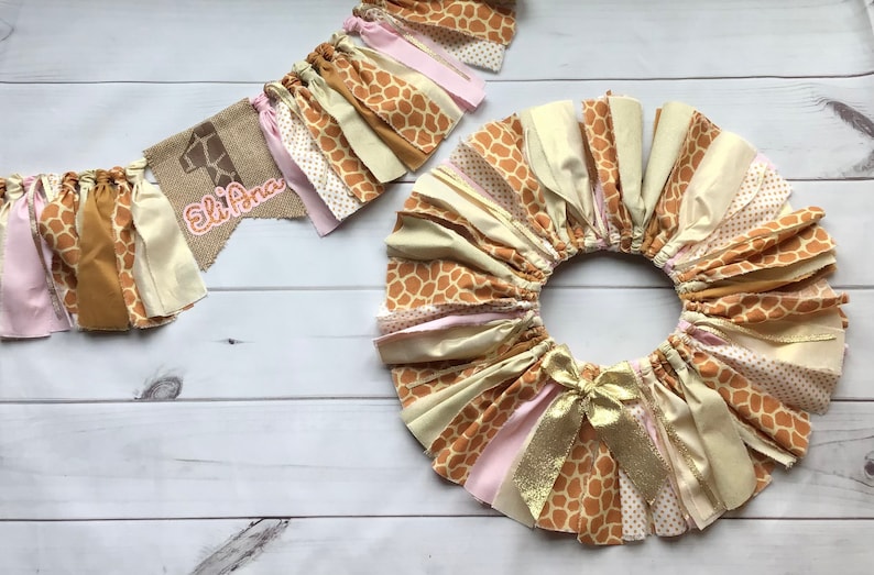 May include: A brown, pink, and gold fabric garland with a giraffe print and the text "Eli Ana" on a burlap banner. The garland is made of multiple strips of fabric with a frayed edge.