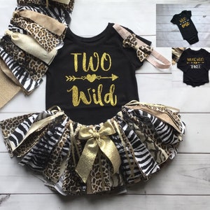 May include: A black and gold two-piece outfit for a baby girl. The top says "Two Wild" with a gold arrow and heart. The skirt is a tulle tutu with leopard, zebra, and beige fabric strips.
