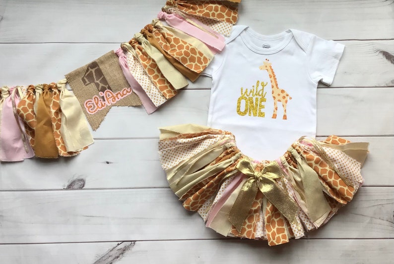 May include: A baby girl's first birthday outfit with a giraffe theme. The outfit includes a white onesie with the words "wild one" in gold glitter, a tulle skirt with pink, gold, and brown fabric strips, and a burlap banner with the name "Eli Ana" and the number "1".