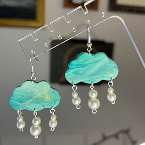 May include: A pair of earrings featuring a blue and green marbled cloud design with white pearl accents. The earrings are hanging from silver hooks.