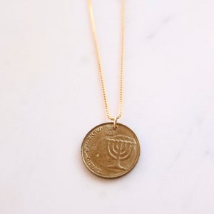 Op de afbeelding: Een gouden ketting met een gouden munt hanger met een menorah ontwerp en de tekst "ישראל" in het Hebreeuws.