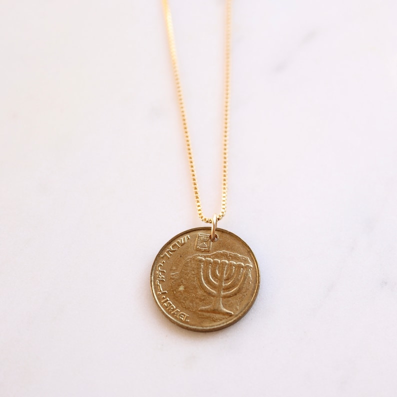 Op de afbeelding: Een gouden ketting met een gouden munt hanger met een menorah ontwerp en de tekst "ישראל" in het Hebreeuws.
