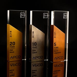 May include: Three clear acrylic awards with a metallic gold, silver, and copper-colored accent. Each award has a different year, number of years, name, and company logo. The awards are displayed on a reflective black surface.
