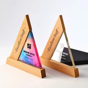 May include: Two wooden triangle-shaped awards with clear and colorful glass inserts. The awards have the text "- Excellent Staff -" and "CREATIVE ARTIST" with a name and the year 2023. The glass insert has a geometric design.