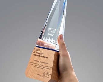Puede incluir: Un premio de vidrio azul y transparente con una base de madera. La parte de vidrio tiene forma de pirámide y tiene el texto "ARTIST YEAR 2022" grabado. La base de madera tiene el texto "NAME SURNAME CERTIFICATE OF APPRECIATION" grabado. La base también tiene el texto "THIS CERTIFICATE IS PRESENTED TO LOREM IPSUM DOLOR SIT AMET, CONSECTETUR" grabado. La base tiene una firma y la palabra "DIRECTOR" grabada.