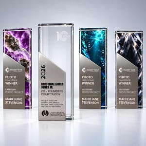 Op de afbeelding: Vier rechthoekige glazen trofeeën met zilveren metalen platen. Drie hebben kleurrijke patronen zoals amethist, blauw water en zwarte diamant met de tekst Master-Land Photo Challenge Winner. Eén heldere trofee toont 10 years, 2026 en tekst.