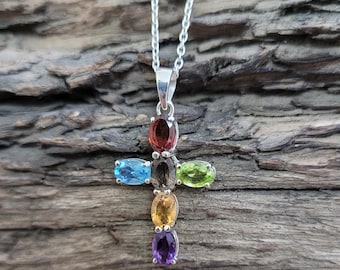 Puede incluir: Un collar de cadena de plata con un colgante de cruz con siete piedras preciosas ovaladas en colores arcoíris. Las piedras preciosas están engastadas en plata y están dispuestas en forma de cruz.