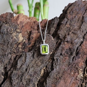 May include: A silver chain necklace with a green gemstone pendant. The pendant is set in a square frame with a smooth, polished finish.