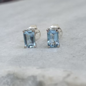 May include: A pair of rectangular, light blue gemstone stud earrings. The gemstones are set in a silver-colored metal. The earrings are displayed on a light gray surface.