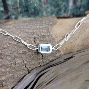 Puede incluir: Un collar de plata con un colgante rectangular de piedra preciosa azul claro. La cadena está hecha de eslabones rectangulares alargados. El collar se exhibe sobre un trozo de madera con un fondo verde borroso.