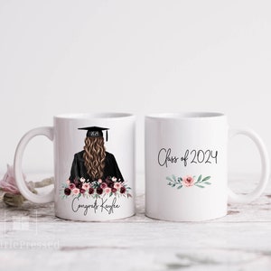 Op de afbeelding: Twee witte keramische mokken. E&eacute;n mok heeft een afstudeerillustratie met een baret, toga en bloemstuk, samen met de tekst "Congrats Kaylee". De andere mok heeft de tekst "Class of 2024" met bloemaccenten.