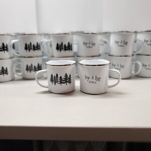 K&ouml;nnte beinhalten: Wei&szlig;e Emaille-Campingbecher mit schwarzen Wald-Silhouetten-Designs und personalisiertem Text. Die Becher haben einen silbernen Rand und einen Griff. Der Text auf einem Becher lautet "Gigi & Issy 27.08.22".
