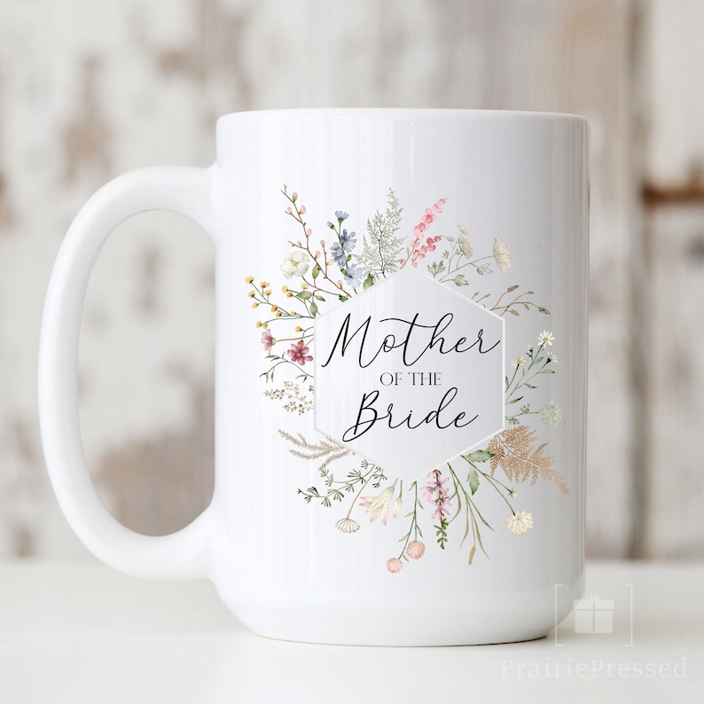 May include: White ceramic mug with a floral design and the text "Mother of the Bride" in a hexagon shape.