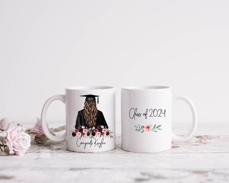 Op de afbeelding: Twee witte keramische mokken. E&eacute;n mok heeft een afstudeerillustratie met een baret, toga en bloemstuk, samen met de tekst "Congrats Kaylee". De andere mok heeft de tekst "Class of 2024" met bloemaccenten.