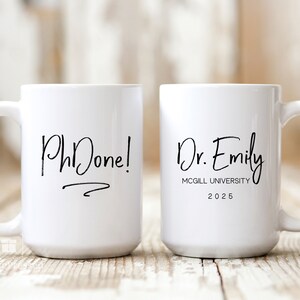 May include: Two white coffee mugs with black text. The mug on the left says "PhDone!" with a line underneath. The mug on the right says "Dr. Emily McGill University 2025".