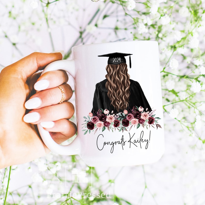 Op de afbeelding: Witte keramische mok met een afstudeerillustratie, inclusief een mortierbordhoed en het jaar 2024. De mok heeft een bloemenontwerp en de tekst "Congrats Kailey!". De mok wordt door een hand vastgehouden.