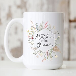May include: White ceramic mug with a floral design and the text "Mother of the Groom" in a hexagon shape.