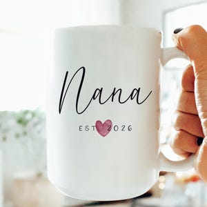 May include: A white ceramic mug featuring the black cursive text Nana, followed by the text EST, a small pink watercolour heart, and the year 2026 in black font. The mug has a smooth finish and a standard handle on the right side for easy gripping.
