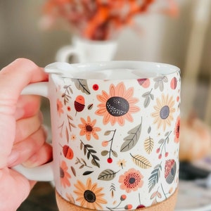 May include: White ceramic mug with a cork base. The mug features a floral pattern with sunflowers, leaves, and berries in shades of orange, yellow, red, and green.
