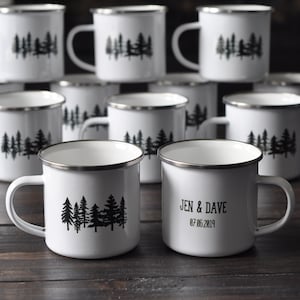 K&ouml;nnte beinhalten: Set aus wei&szlig;en Emailletassen mit schwarzen Wald-Silhouetten-Designs. Einige Tassen haben den Text "Jen & Dave 07.06.2019" aufgedruckt.