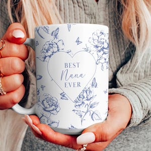 May include: A white ceramic mug featuring a blue floral and butterfly pattern. In the centre, a large heart outline contains the text BEST NANA EVER in blue lettering. The mug has a curved handle and a smooth finish, displaying a classic design.
