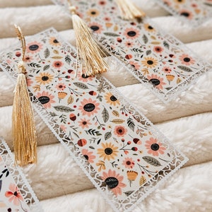 May include: A set of white, laser-cut bookmarks with a floral design featuring pink, yellow, and orange flowers. Each bookmark has a gold tassel.