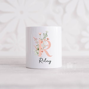 Peut inclure: Une tasse en céramique blanche avec un motif floral rose et la lettre "R" en rose. Le nom "Riley" est écrit en écriture noire sous le motif floral.
