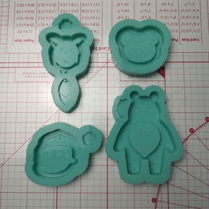 Puede incluir: Tres moldes de silicona azul turquesa para hacer manualidades de resina. Los moldes tienen forma de oso, oso con pajarita y persona con sombrero. Los moldes están sobre un fondo de cuadrícula blanca.