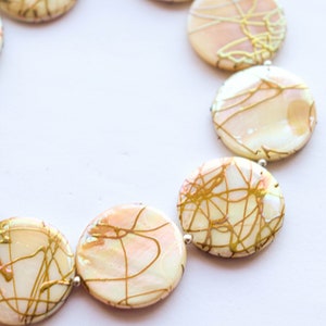 May include: A necklace made of round, white, iridescent shell beads with gold painted designs. The beads are strung on a silver chain.