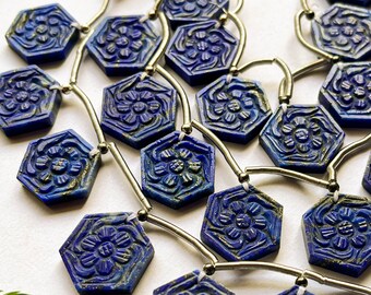 May include: Hexagonal blue lapis lazuli beads with carved floral designs. The beads are strung together with silver-colored wire, and a white tassel is visible. The beads are a deep blue color with gold accents.