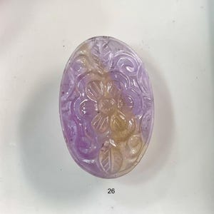 Puede incluir: Un colgante de piedra ovalada tallada con un dise&ntilde;o floral. La piedra es una mezcla de morado y amarillo.
