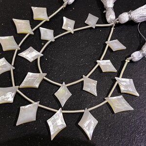 May include: Strands of iridescent, diamond-shaped mother-of-pearl beads. Each bead is strung on a silver wire, with a tassel at the end. The beads have a pearlescent sheen, reflecting light. Ideal for jewelry making.
