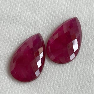May include: Two loose, pear-shaped rubies with a faceted surface. The gemstones are a deep, saturated magenta-red color, displaying a polished finish that reflects light across their geometric, multi-faceted tops and smooth, rounded edges.