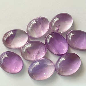 May include: A collection of eight oval-shaped amethyst gemstones. The stones display a gradient of colors, ranging from pale lavender to deep purple. Each stone has a polished, glossy surface, reflecting light. The gemstones are arranged on a white surface.