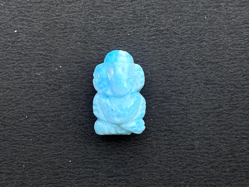 K&ouml;nnte beinhalten: Eine hellblaue, geschnitzte Figur einer sitzenden Gottheit, m&ouml;glicherweise Ganesha, mit gro&szlig;en Ohren und einem R&uuml;ssel. Die Figur besteht aus hellblauem Stein und ist vor einem dunklen Hintergrund platziert.