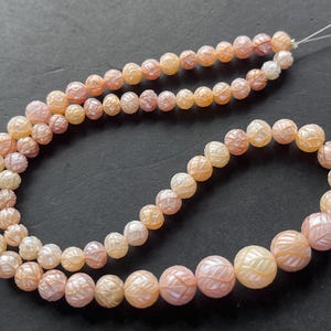 May include: A strand of carved, peach-colored beads. The beads are round with a detailed, etched pattern. The beads vary in size, with the largest beads at the bottom. The beads are strung on a thin, white string.
