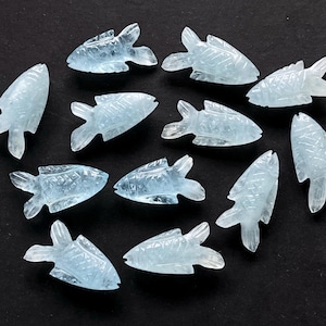 May include: A collection of aquamarine-colored, carved fish figurines. Each fish has detailed fins and scales, and they are arranged on a dark surface. The translucent quality of the stone gives them a delicate appearance.