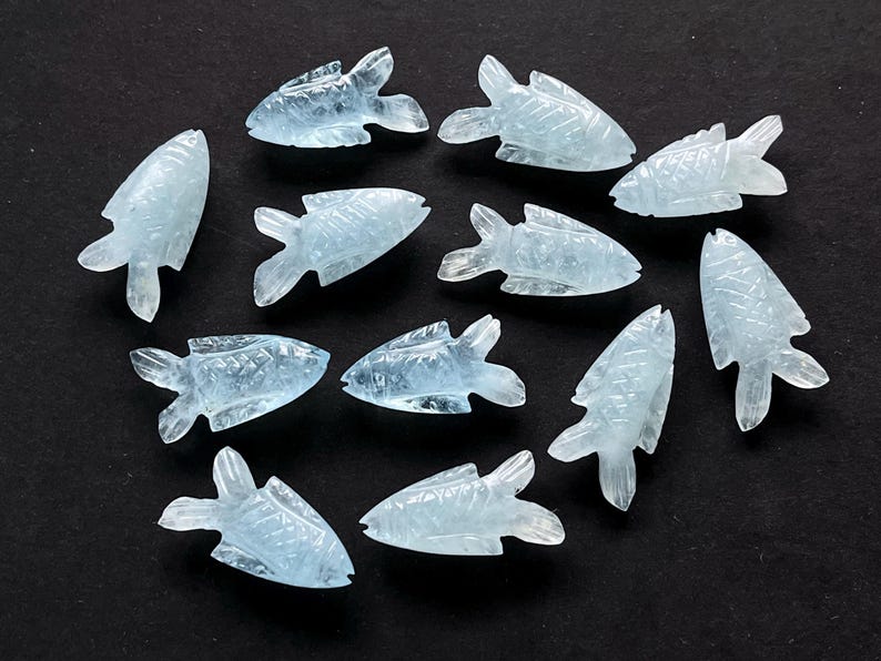 May include: A collection of aquamarine-colored, carved fish figurines. Each fish has detailed fins and scales, and they are arranged on a dark surface. The translucent quality of the stone gives them a delicate appearance.