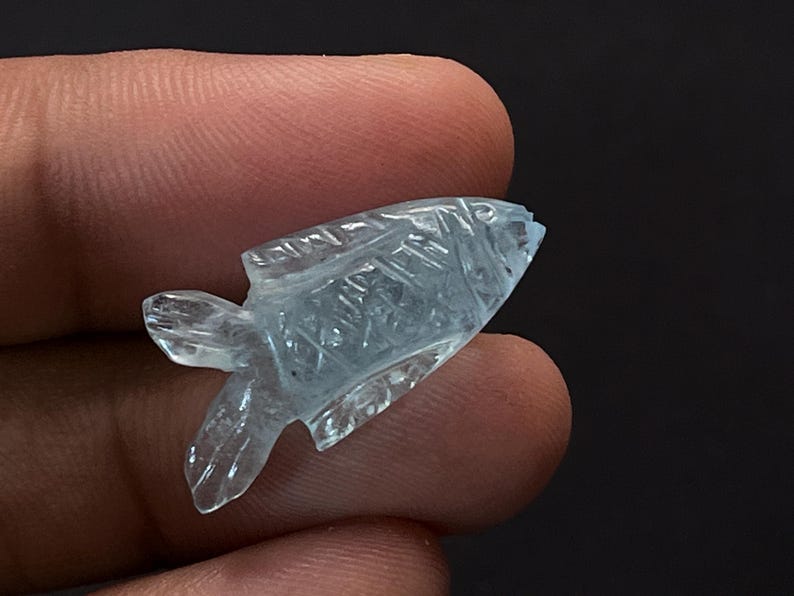 May include: A carved, light blue gemstone fish figurine. The fish has detailed etched lines and fins. The gemstone is held between fingers against a dark background. The fish is a decorative item.