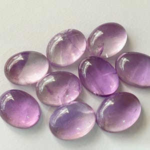 May include: A collection of eight oval-shaped amethyst gemstones. The stones display a gradient of colors, from pale lavender to deep purple. Each stone has a polished, glossy surface, reflecting light. The gemstones are arranged on a white surface.