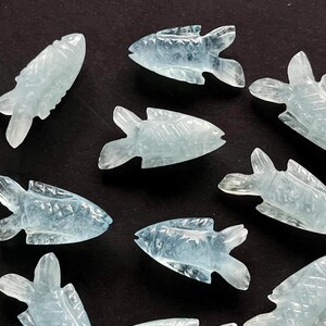 May include: A collection of light blue, carved gemstone fish figurines. Each fish has detailed fins and a carved body, with a slightly translucent appearance. The figurines are arranged on a dark surface.